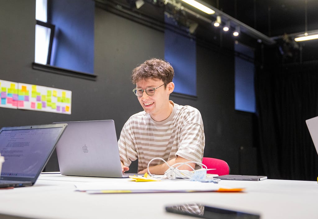 A young man with glasses and light brown hair sits down in front of a laptop and is smiling. He is a room with dark walls and sticky notes appear from the right side edge of the photo.