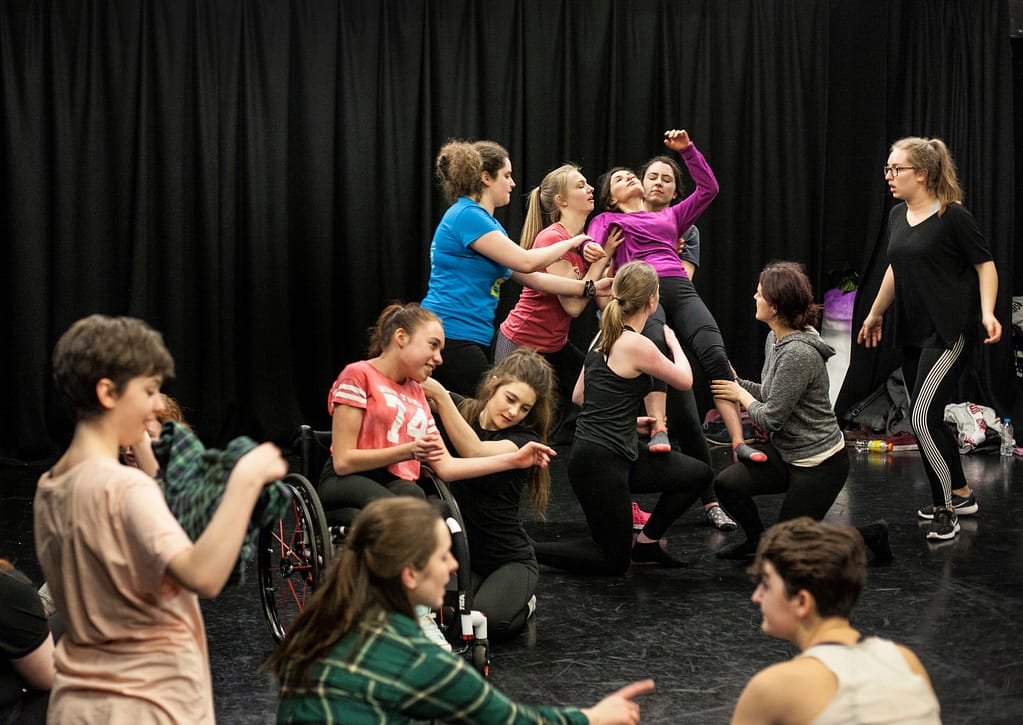 A group of performers cover the floor of a black box theatre in various positions, some are kneeling, some are standing, some are lying down, some are leaning against one another. There is a black curtain in the background