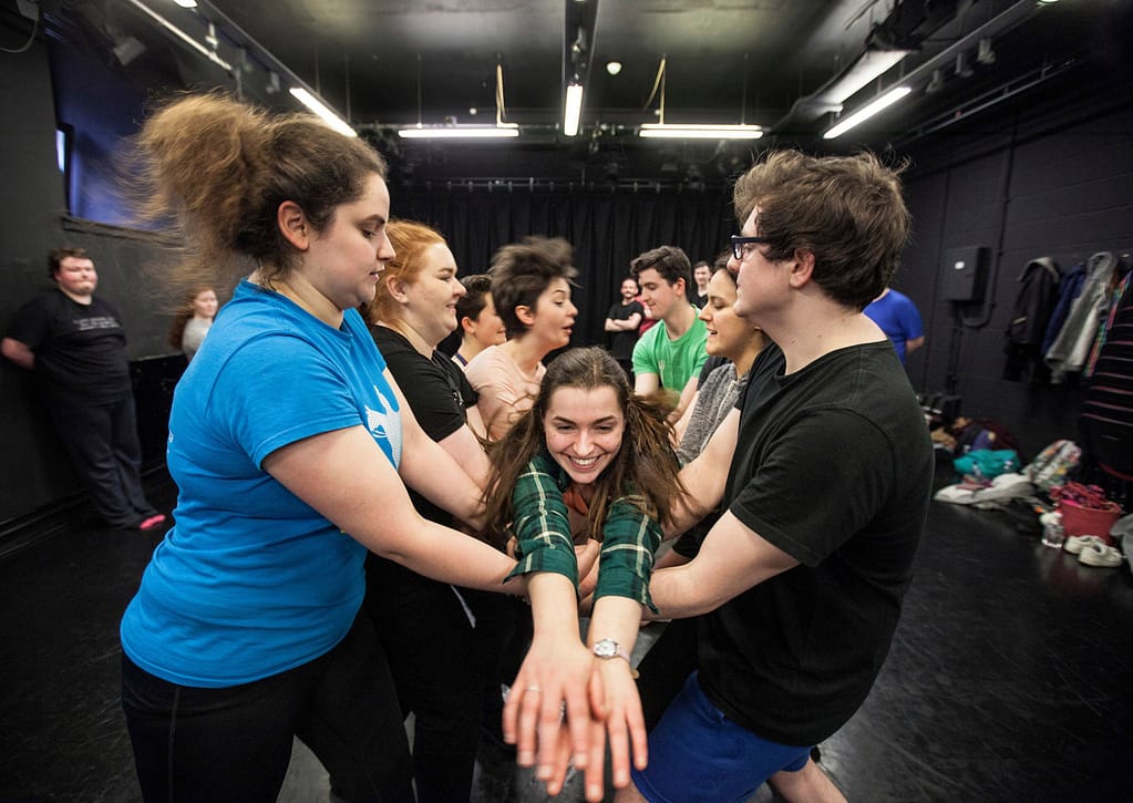 Two lines of young people face one another with hands clasped in the centre, catching a young person jumping between them