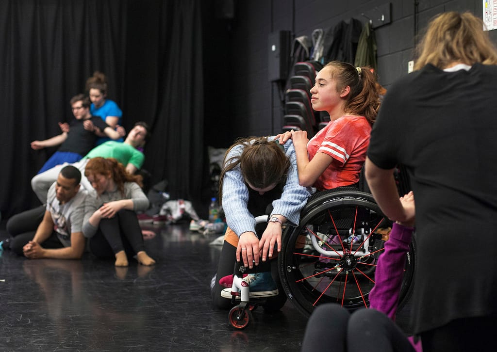 Various performers lie, kneel, sit or stand on the floor of a black box theatre
