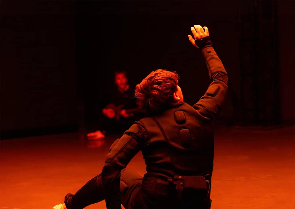 A performer with short hair is dressed in a black padded suit with wires protruding from the back and hip. They are seated, with one arm on the ground and one arm reaching into the sky. There is a strong orange glow in the image