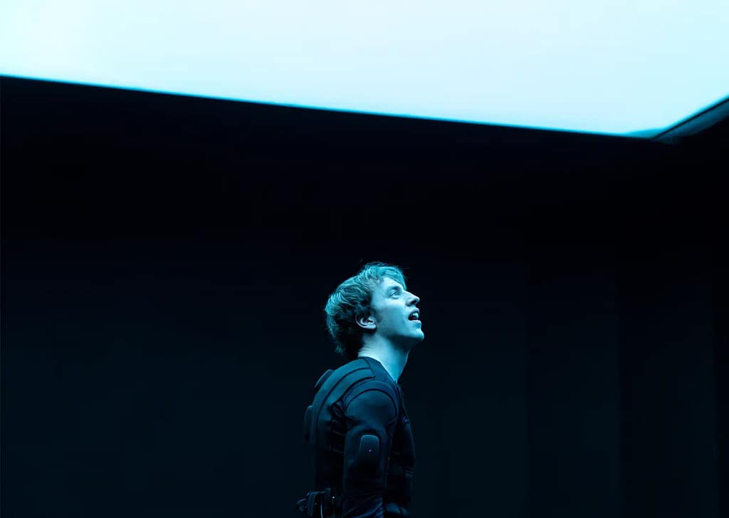 A performer with short hair is dressed in a black padded suit with wires protruding from the back looks up to a bright blue/white light shining above them.