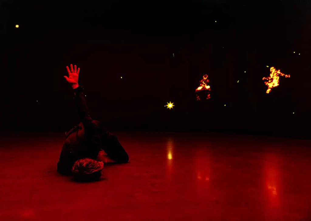A performer with short hair is dressed in a black padded suit with wires protruding leans in a convoluted position on the floor, face against the ground with one arm up, fingers spread into the air. They are in front of a large screen that shows stars and human like figures made from fire. The image has an intense red glow to it