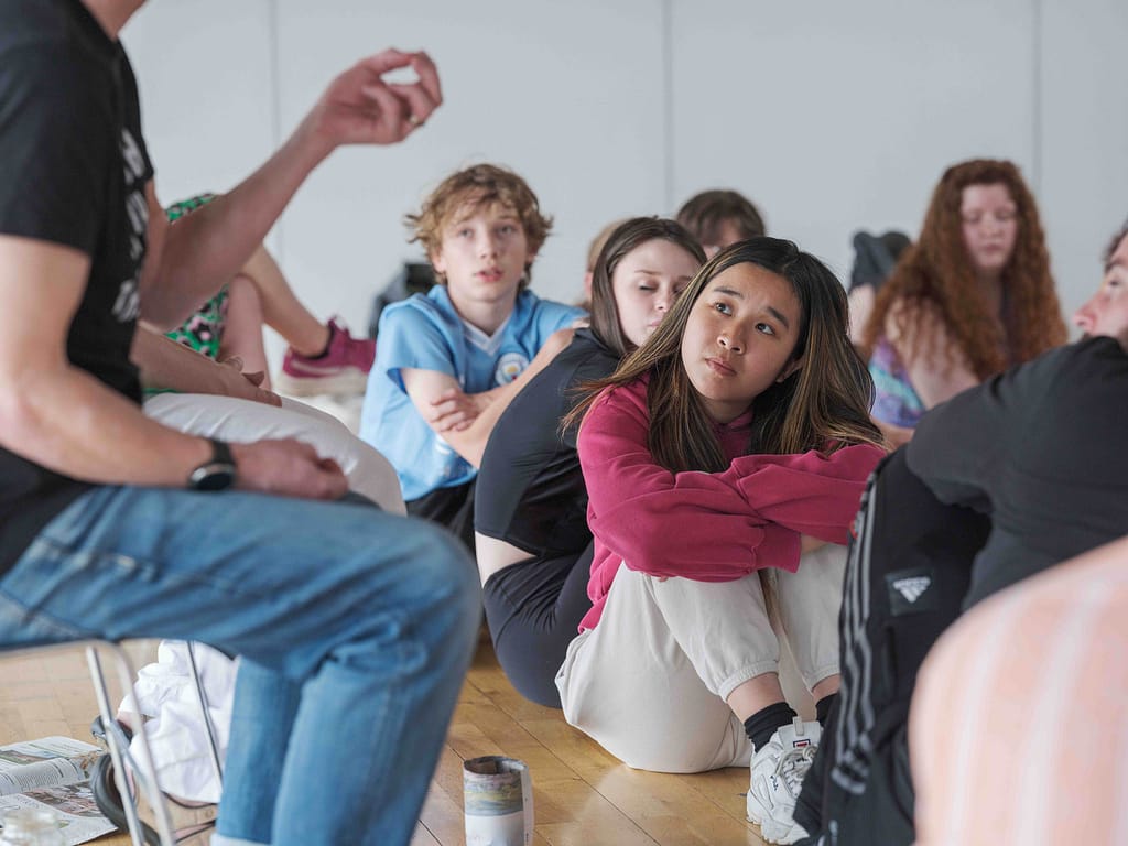 A group of young people sit on a wooden floor. The person in the centre of the image is wearing a pink jumper and hugs their knees to their chest, looking up to a person sat on a chair just out of shot to the left.