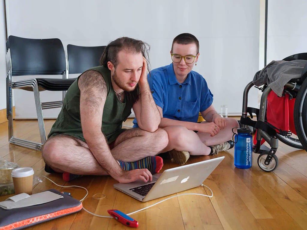 Two people sit cross legged on the floor looking at a laptop screen in front of them placed on a wooden floor. They are surrounded by different objects including a paper coffee cup, a laptop case, a stanley knife, a blue water bottle and a wheelchair. There are also some black stacked chairs behind them.