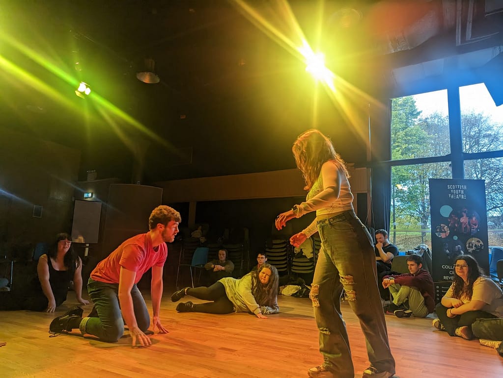 A young performer leans forward with her side to the camera, face covered by her hair. She faces another young person on their knees with their arms outstretched in front of them. The two figures are surrounded by young people sitting cross-legged on a wooden floor. The scene is washed in bright yellow light.