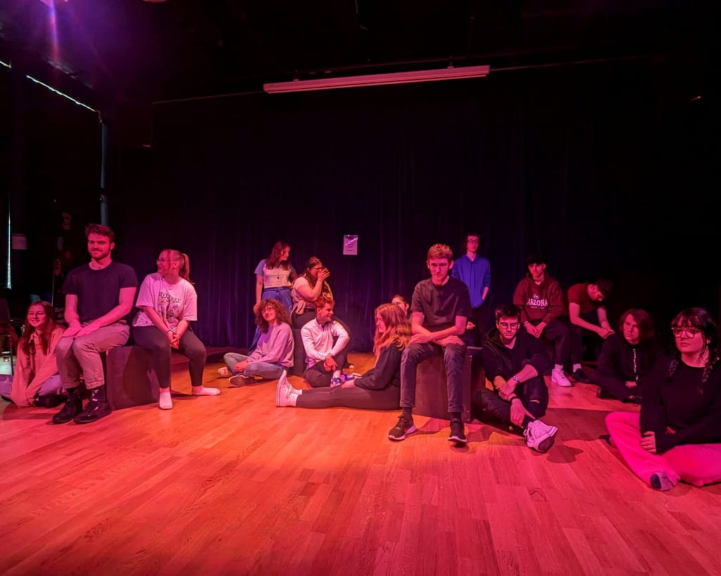 A group of young people all gather on a wooden floor in various positions. Some sit on wooden boxes, some sit crossed legged on the floor, some are standing. All have serious looks on their face. A black curtain is behind them in the background and the scene is awash in a strong pink hue.