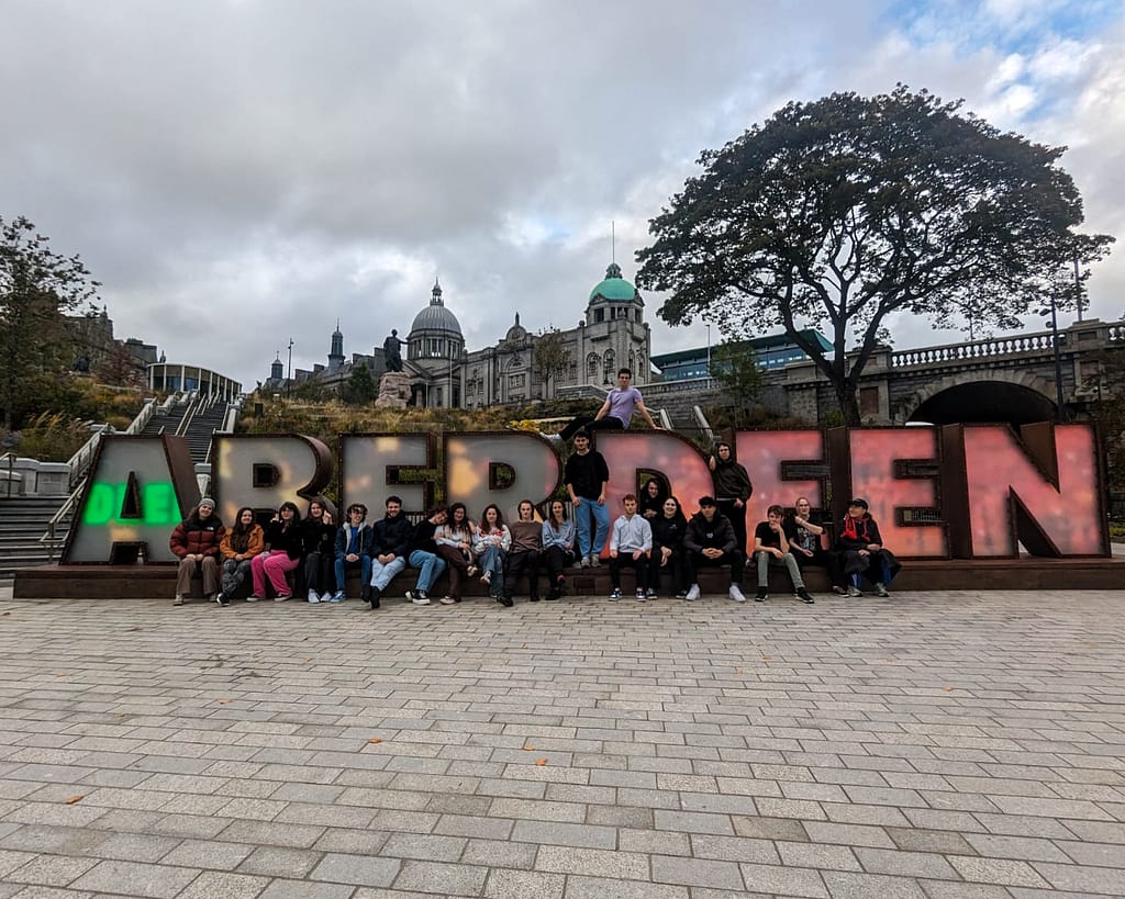 A group of 21 people sit and stand on a large letters spelling out 'ABERDEEN' on a stone paved ground. One person is lying across the top of the 'R' and 'D'. There is a tree and bridge in the background, with stairs leading off the left side.