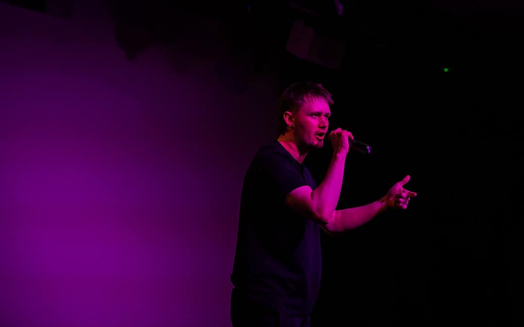 Performer stands in a pink light speaking out into a microphone. They have short hair and wear all black. 