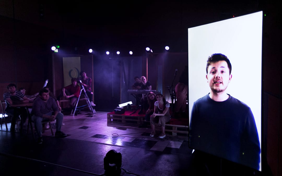 A digital screen is in the foreground displaying a young man in a dark jumper smiling and looking out to the audience. A group of performers on a purple lit stage are in the background, sitting on various props, watching the screen.
