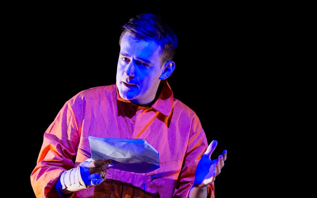 'MAKING SPACE' – A performer lies on a raised platform in partially blue light. They wear orange overalls and hold a script in their hand.