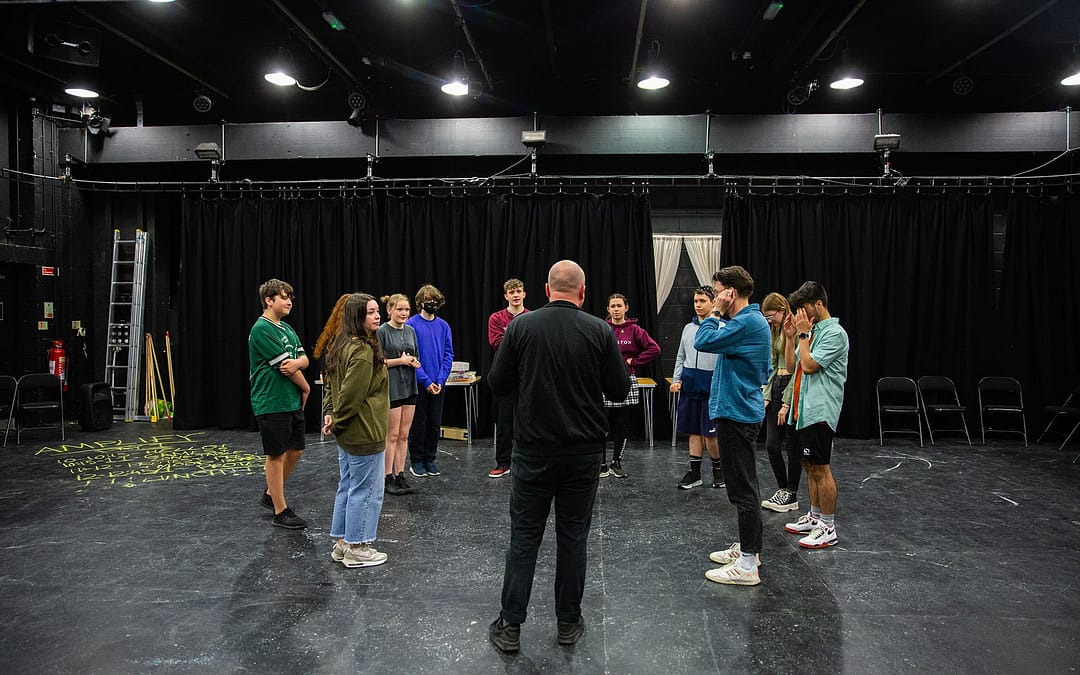 A group stand in a circle facing one another, standing on a black floor in a black box theatre, with dark curtains in the background. There is chalk writing on the floor to the left of the image.