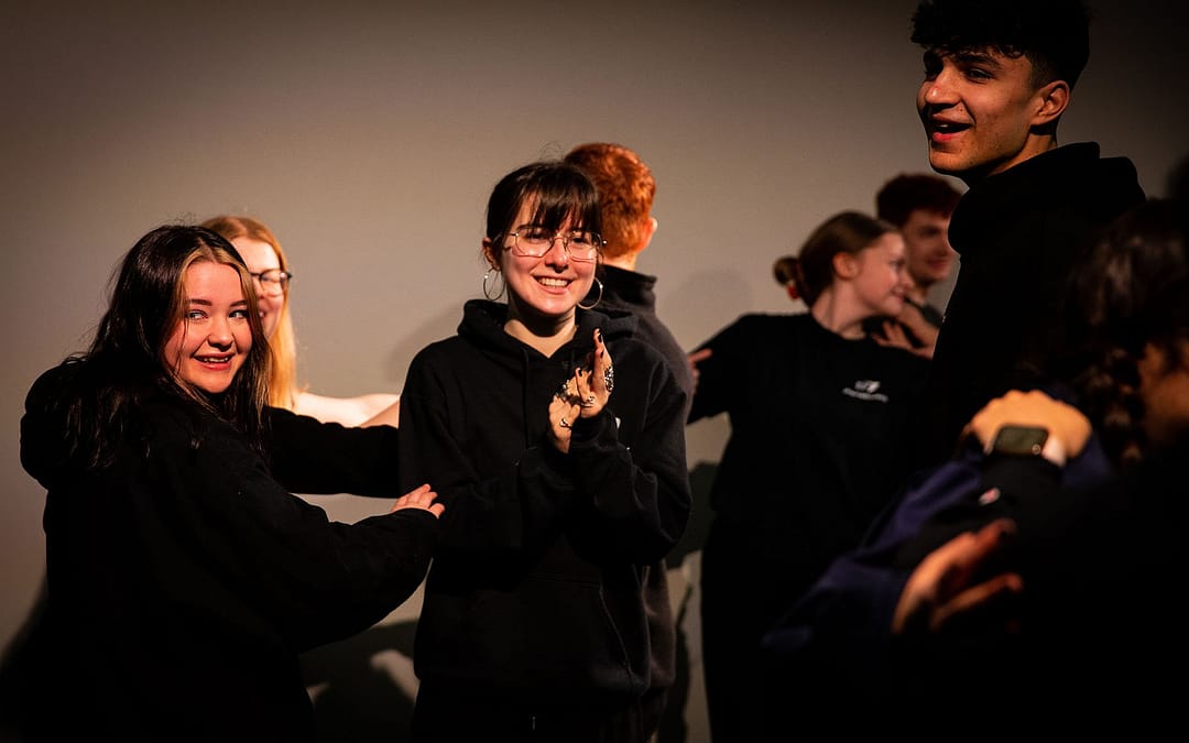 A group of young people dressed all in black look to the left and right of camera. They all smile and the person in the middle is clapping. More figures dressed in black are dancing and moving in the background.