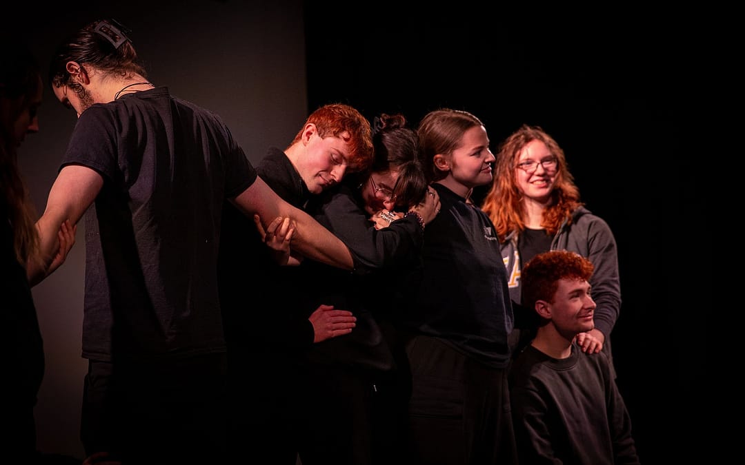 A group of young people dressed in dark clothing huddle together, grasping on another's arms and shoulders. They have a mixture of happy and content expressions on their faces