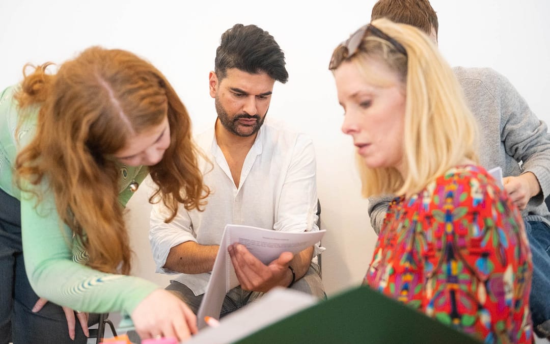 Two women (one with ginger hair, one with blonde) in the foreground look at a script and point to it. Two men in the background (one with black hair, one with brown) look at separate scripts and look inquisitively at them. There is a white wall behind them.
