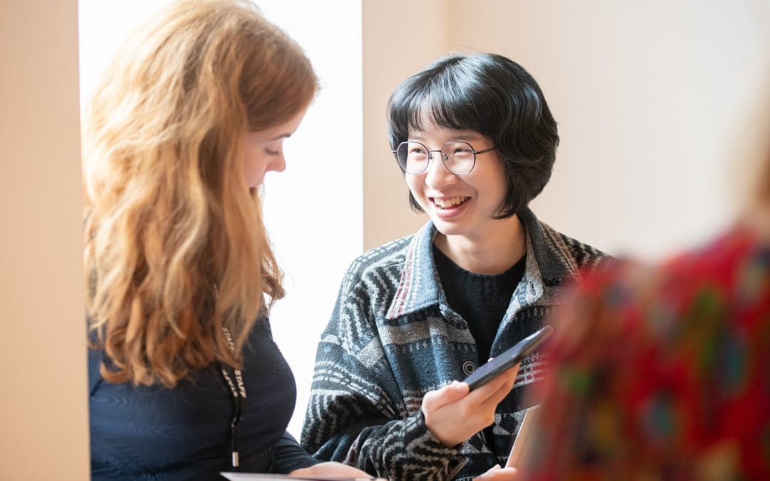 A woman with long ginger hair looks and smiles at young person with short black hair and thin-framed glasses who is showing her their phone. They both wear dark clothes with their backs to a window where sunshine is bleeding through.