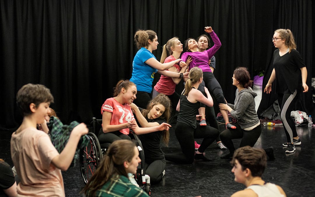 An ensemble group of young people take various dramatic positions on a black floor; sitting, kneeling and standing. One person wearing a purple jumper collapses backwards while 5 people catch and support her. There is a black curtain in the background.