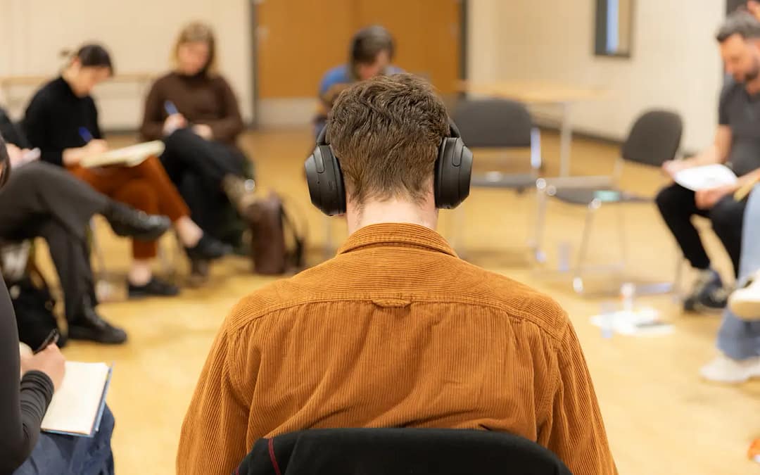 A young man with a mustard coloured corduroy shirt on has his back to the camera. He is seated, looking down with headphones on. Blurred in the background other people sit in chairs looking down writing on notepads on their laps.