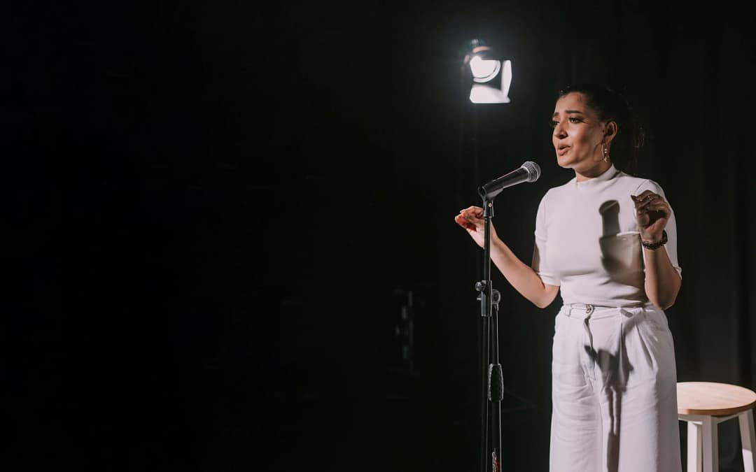 A young woman dressed all in white stands in front of a microphone, lit by a single spotlight in a black box theatre, with a wooden stool behind her, gesturing into the mic