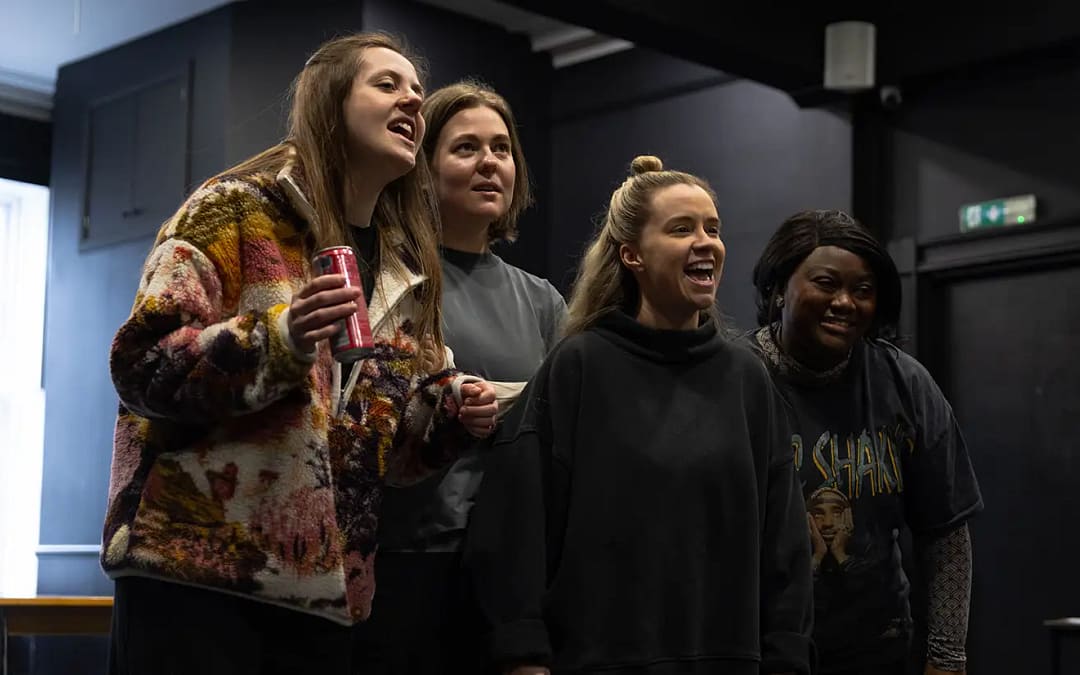4 young women wearing dark jumpers and tops stand in a line looking to the side of the camera. They have a range of expressions on their faces, from smiling, to laughing, to wonder. The woman on the far left holds a can in her left hand