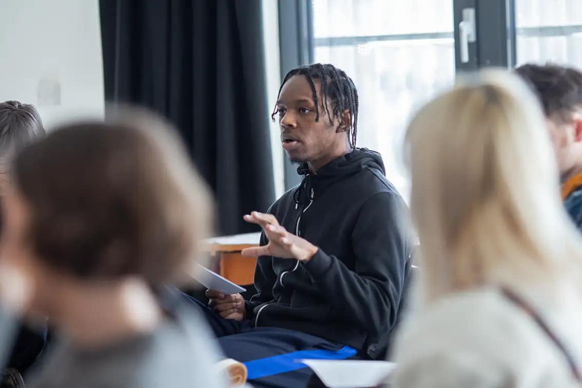 A young black man in a black hoodie sits on a chair looking outwards. He holds a sheet of paper/script in one hand while gesturing with slightly clawed fingers with the other