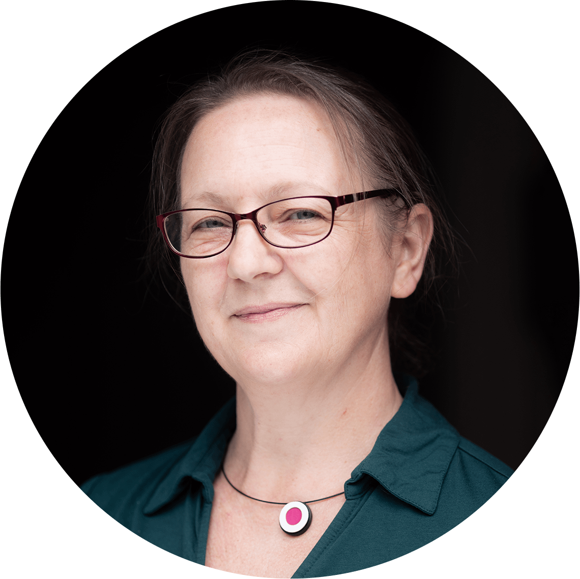 A woman with a dark green blouse, dark rimmed glasses and a black, white and pink minimalist necklace smirks at the camera