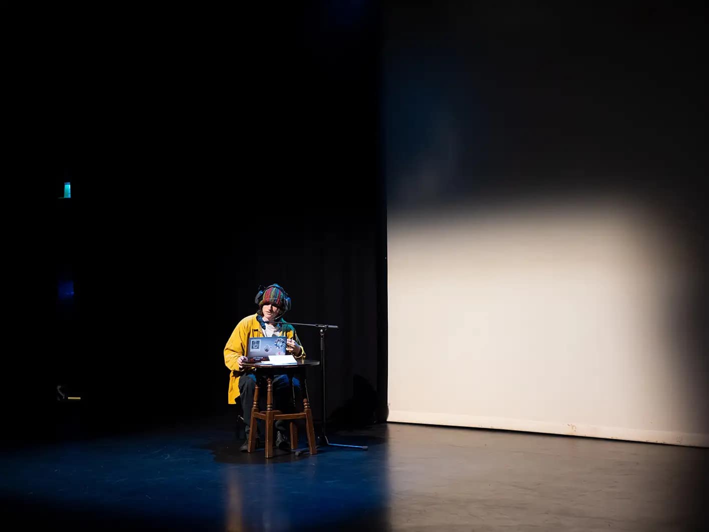 A young person dressed in a bright yellow overshirt and multicoloured beanie, wearing headphones, sits isolated at the side of the stage, perched on a small stool. They sit in front of a microphone and a laptop opened in front of them on a small table. They are highlighted by a bright spotlight, with a screen to the side of them.