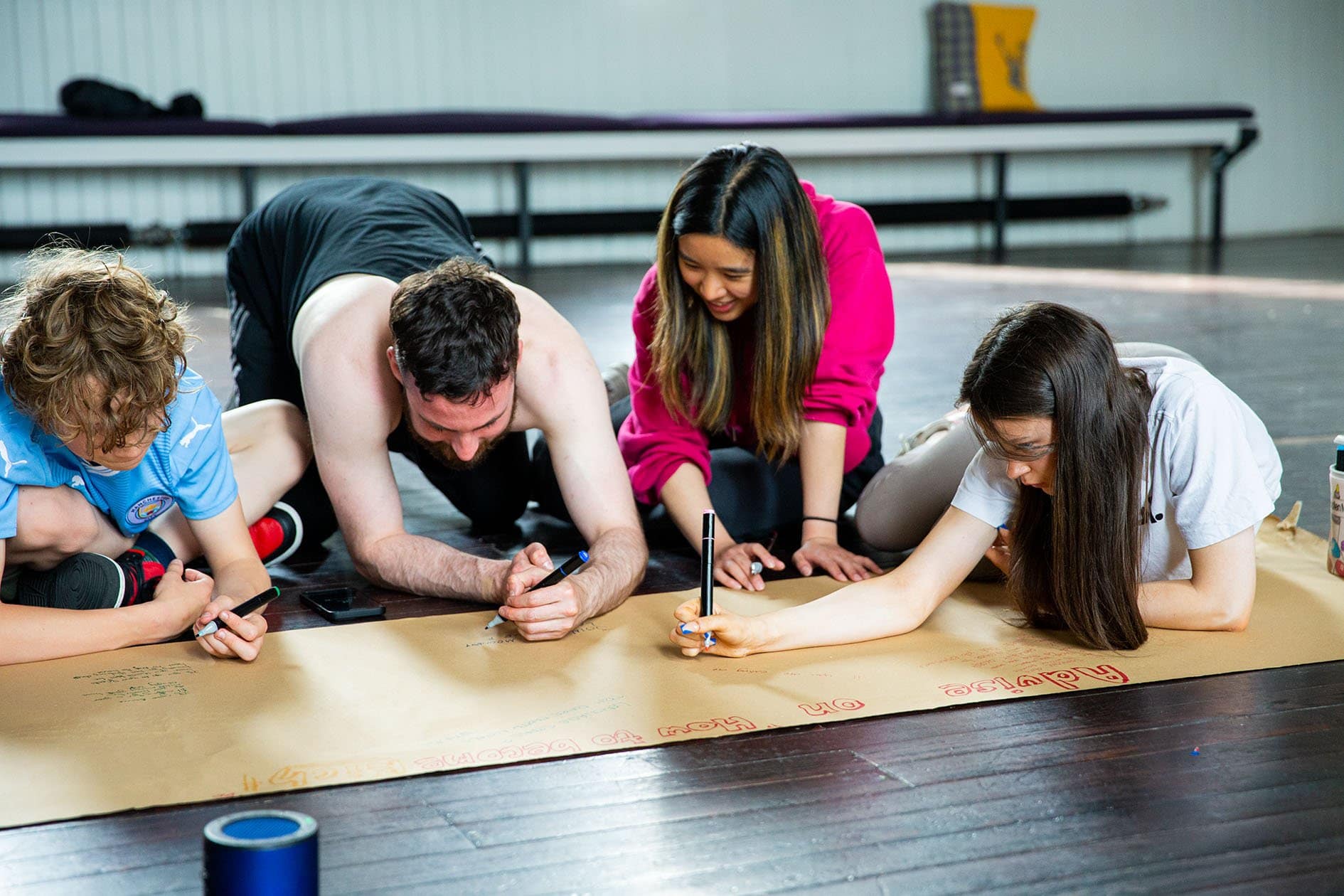 An artist and three young people kneel on a wooden floor with a scroll of brown kraft paper in front of them. They smile while writing on the scroll with different coloured pen markers.