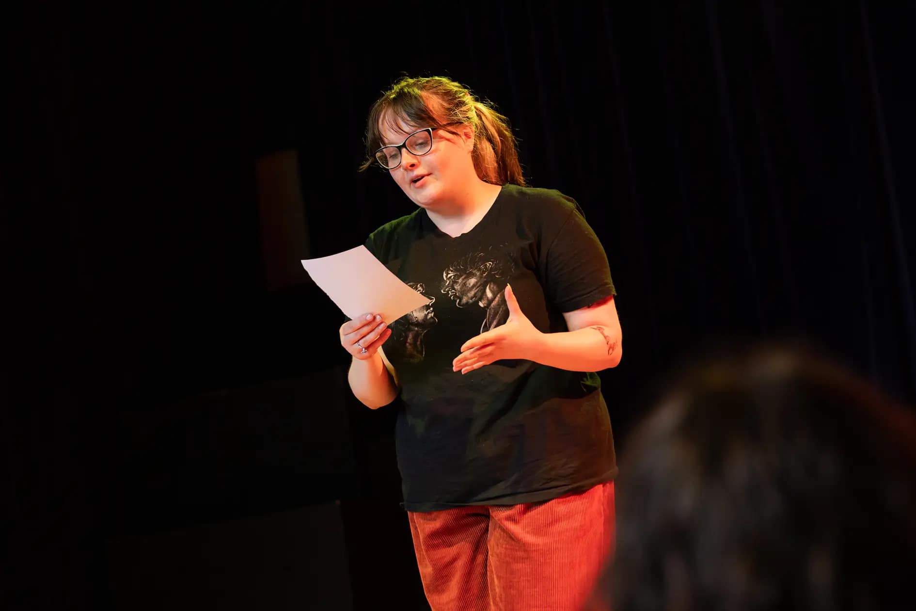 A young person in a black t-shirt and rust coloured corduroy trousers stands alone, holding a script in their right hand, outstretching their left hand in front of them. They look inquisitively at the sheet of paper when performing it