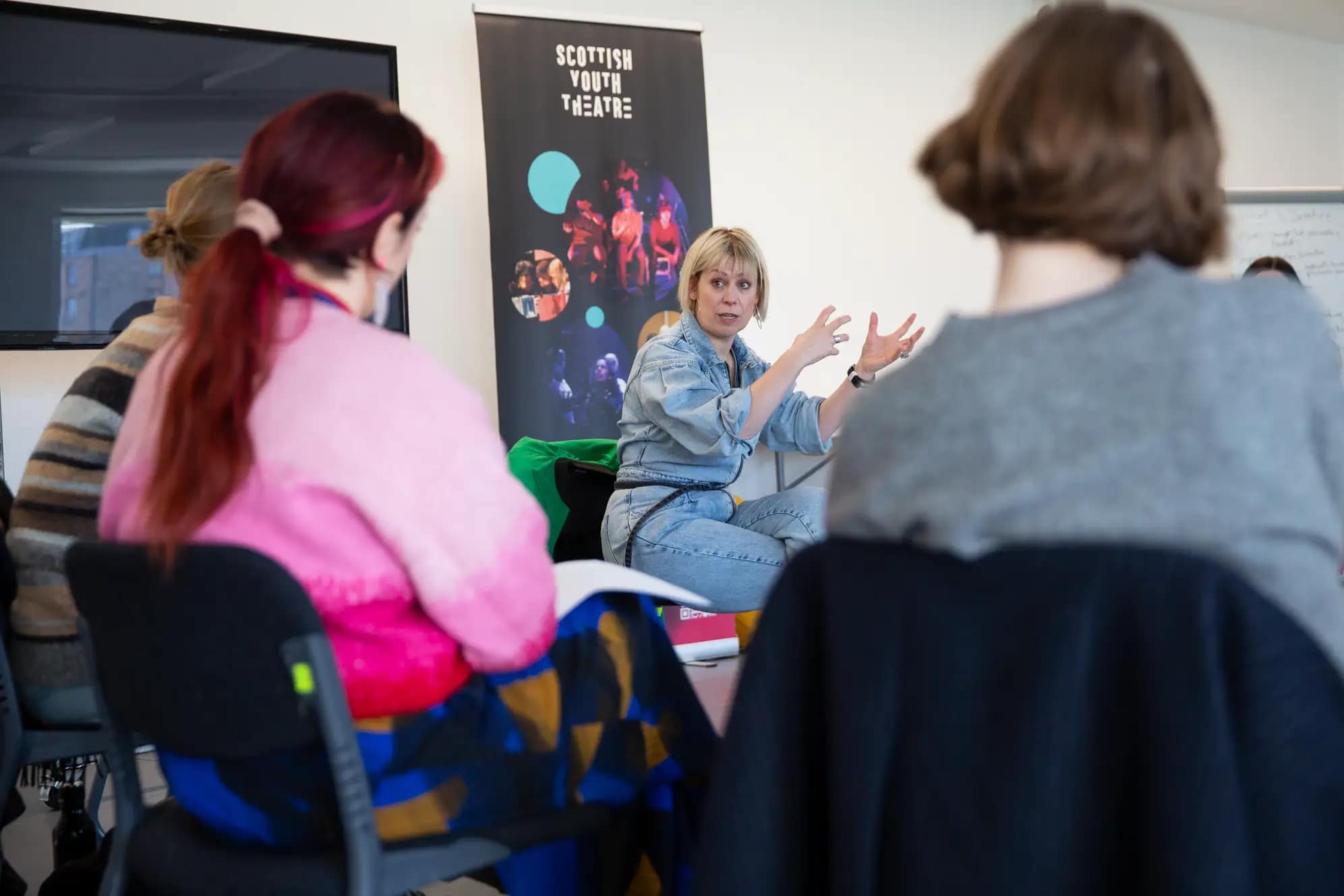 A group of people sat huddled in chairs look intently towards a woman with short blonde hair, wearing a blue denim jumpsuit. Her hands are raised in the air, gesturing with clenched fingers
