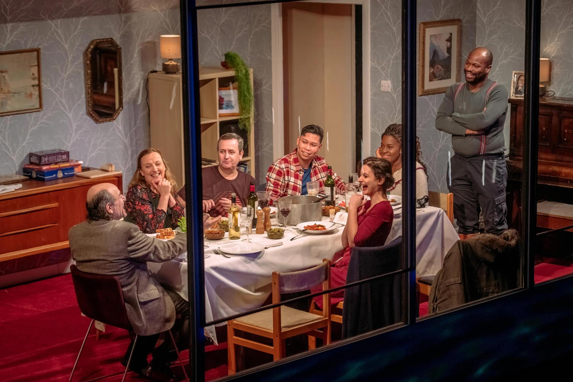 A diverse range of people are on stage behind a glass window. There is a mix of ages and ethnicities present around the table. Their attention is focuses at a light-skinned older man who appears to be telling a funny story.