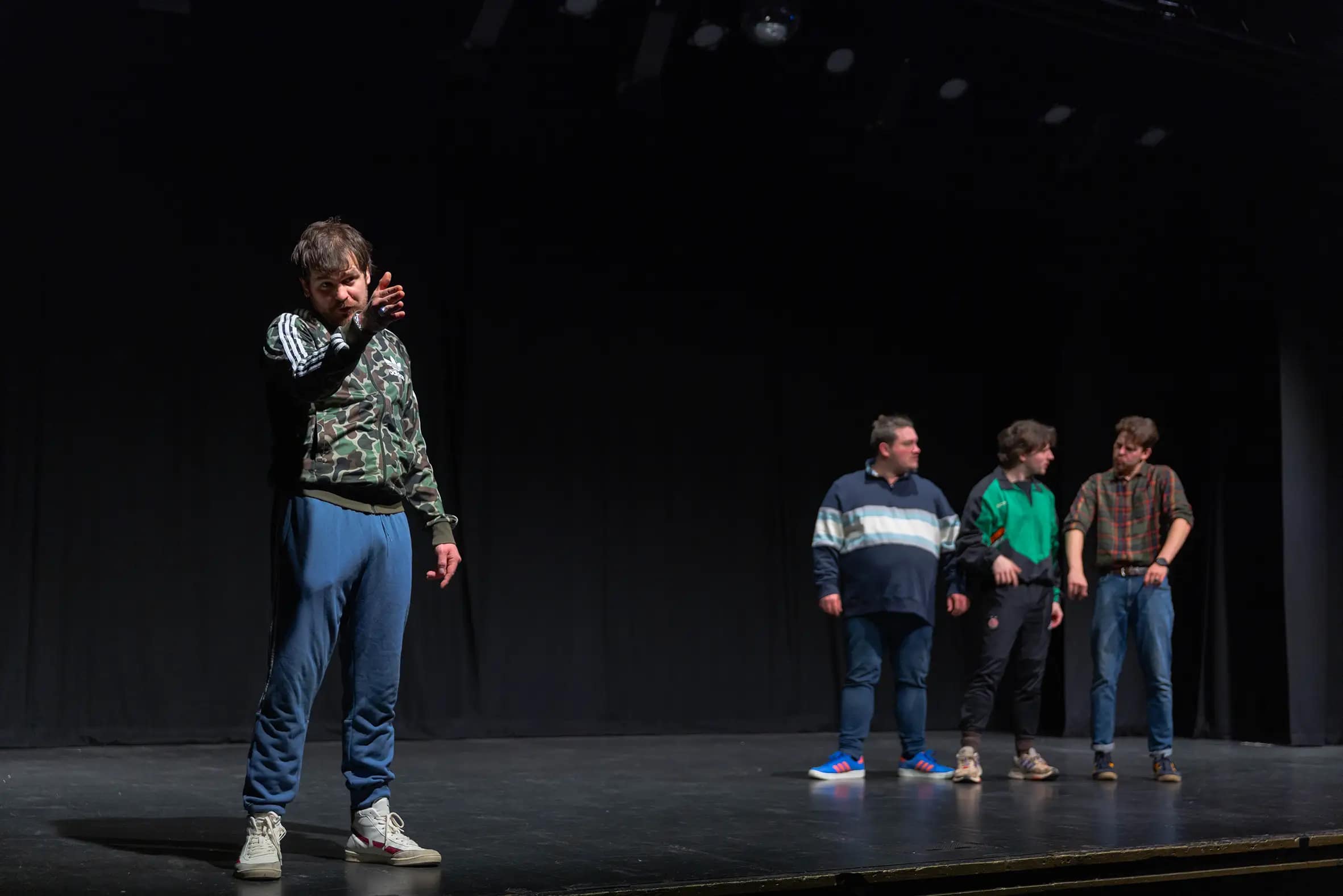 4 young men, dressed in casual sports gear, stand huddled on a dark stage. One wearing a camo patterned green adidas track top and navy blue joggies, stands at the front, gesturing emphatically out towards the audience