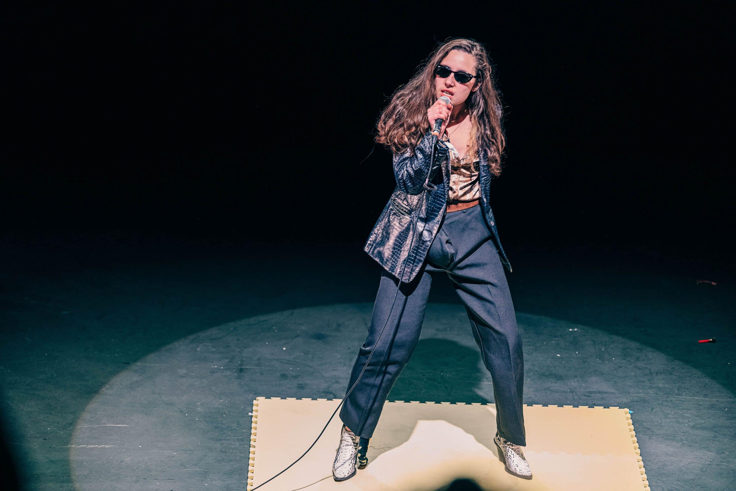 A young woman dressed in a black snakeskin effect jacket, black trousers and snakeskin effect cuban heeled boots poses in the middle of a stage, holding a microphone in one hand, other hand behind their back, with a bright white spotlight shining on them