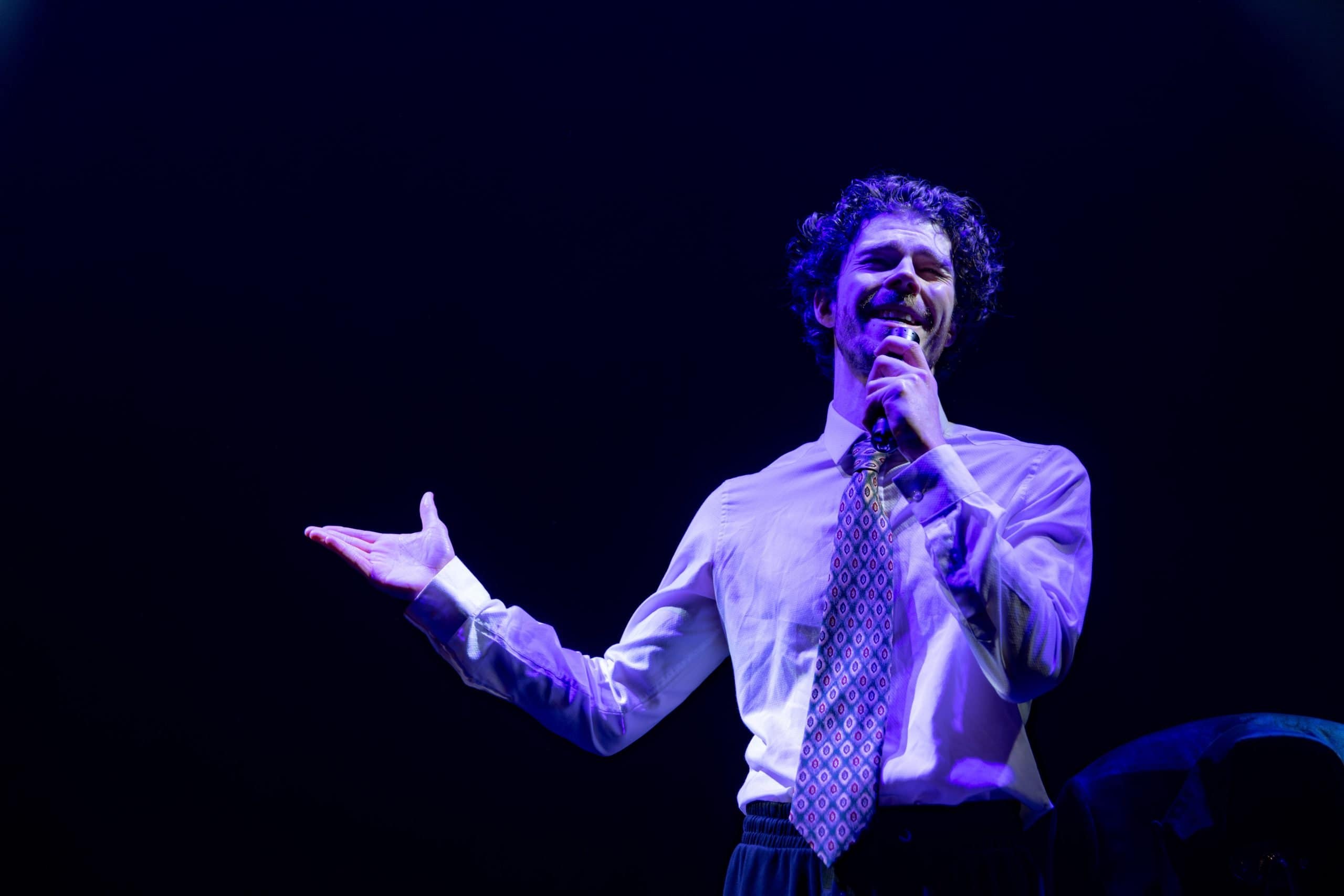 A performer stands on stage, bathed in blue light and speaking into a microphone. They wear a shirt and tie.