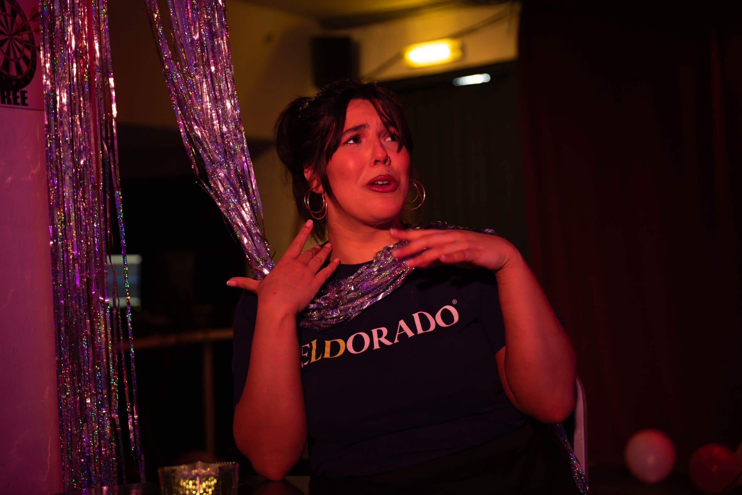 A performer sits in a pub-like setting with tinsel wrapped around their neck, enthusiastically tell a story.
