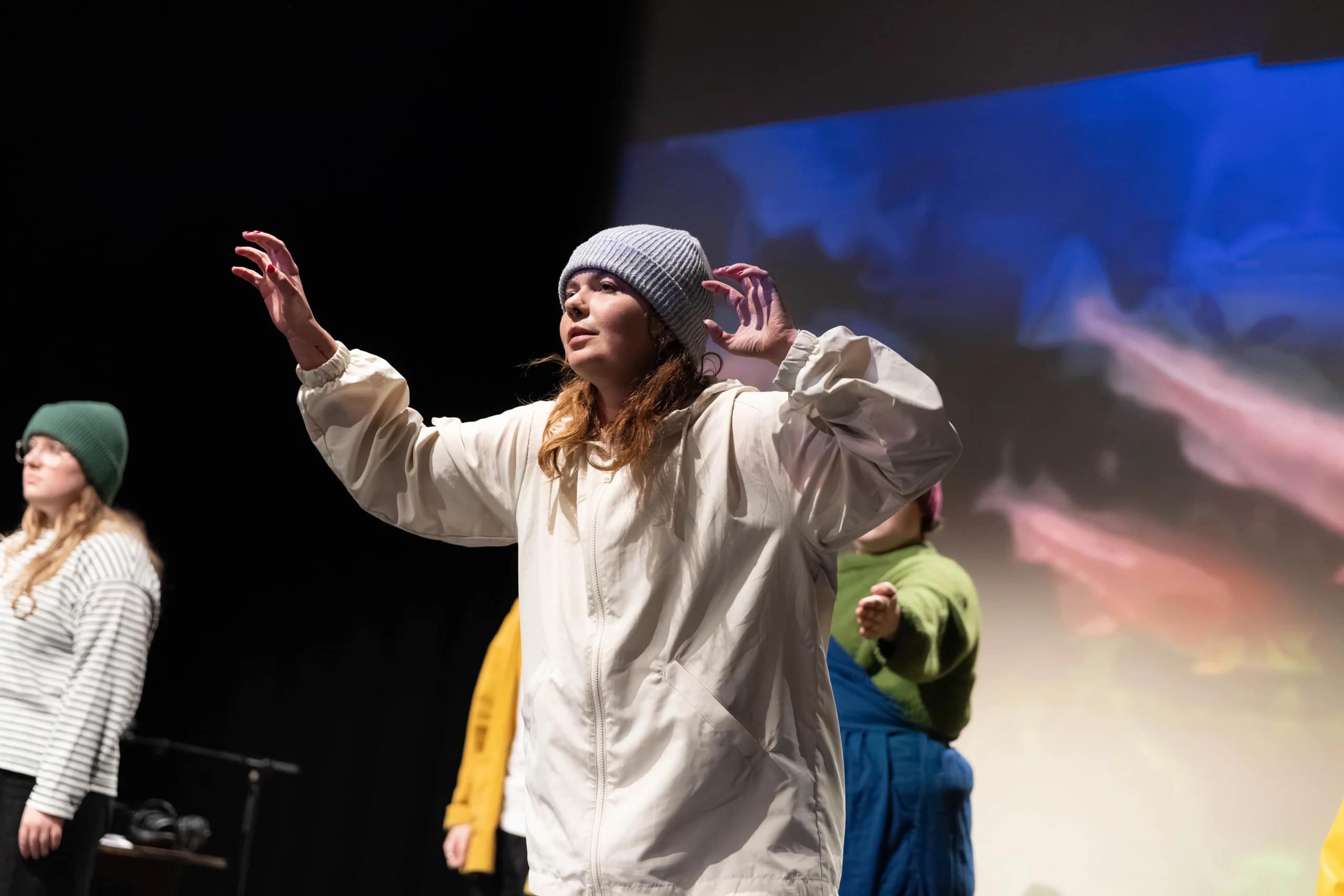 A performer, dressed in a grey beanie and beige coloured cagoule, holding their arms up pensively, clawing their fingers, with other performers in the background.