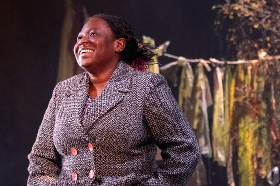 An actor with dark skin and long black hair tied back smiles on a stage. They wear a grey blazer and a washing line hangs behind them.