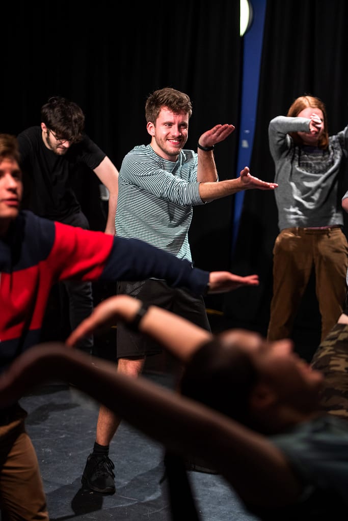In the centre is a young performer gesturing with both arms outstretched. They wear a black and white striped long-sleeved top. Around them are other performers in various positions with flailing arms.