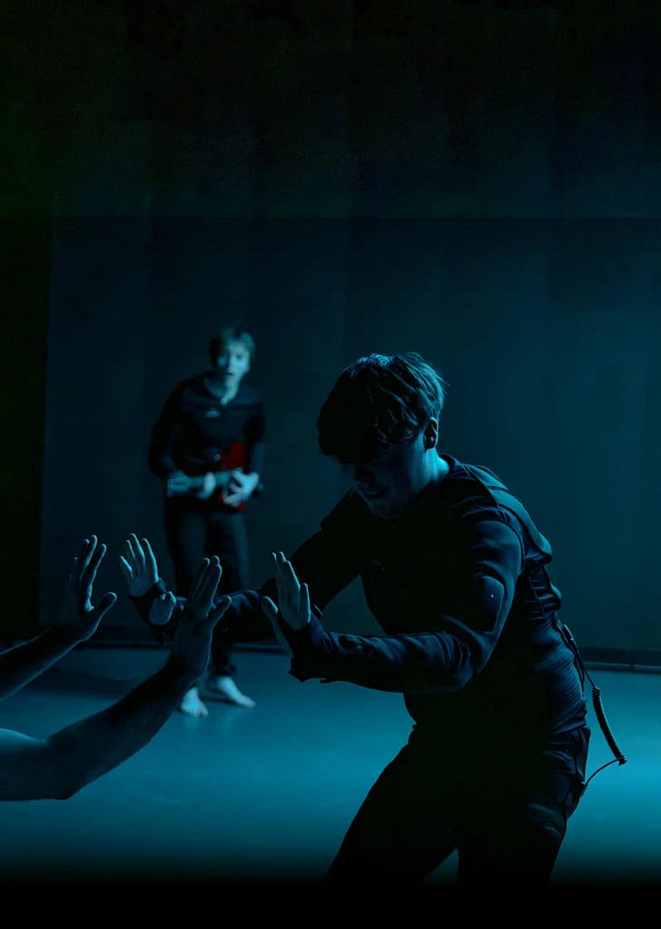 A performer with short hair is dressed in a black padded suit with wires protruding from the back and hip. They face the audience and hold their hands out in front of them, where they are met by another set of hands. Another performer stands in the back holding a guitar, watching intently. There is a strong blue hue to the image.