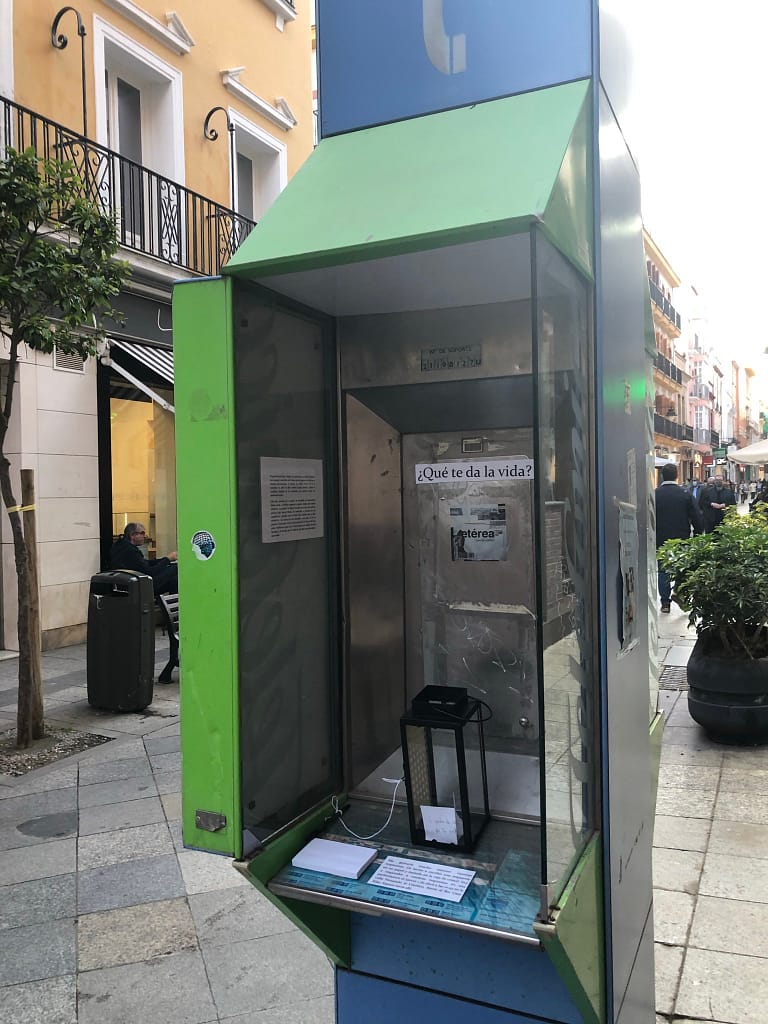A metal green and blue phonebox in the middle of a street displays some small paper plaques and a candle holder inside. The legible text reads "QUÉ TE DA LA VIDA?"