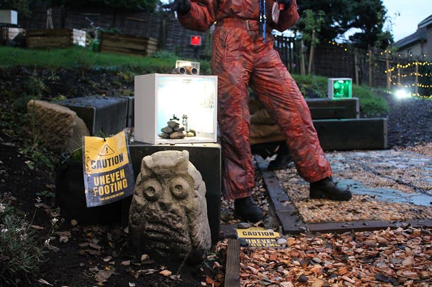 The lower half of a person dressed in a red jumpsuit and black boots stand next to a plinth and a stone sculpture of an owl. Inside the plinth is a small pile of pebbles. There is a yellow and black sign next to the plinth that reads "CAUTION UNEVEN FOOTING"