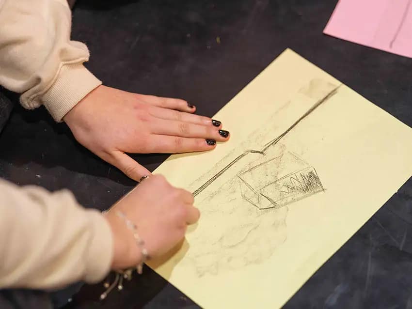 Two hands with black painted fingernails hold a yellow sheet of paper firmly on the floor. There is a sketch of a box and some dynamic lines on the sheet. They continue to draw on the piece of paper with some charcoal. There is a piece of pink-coloured paper in the background.