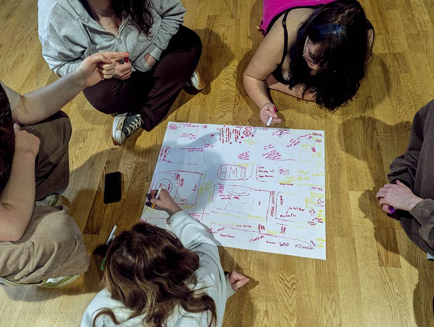 A group of 5 young people sit and lie on a wooden floor around a large sheet of white paper. The paper has yellow and red writing and scribbles on it. Two of the young people hold yellow and red pens in their hands.