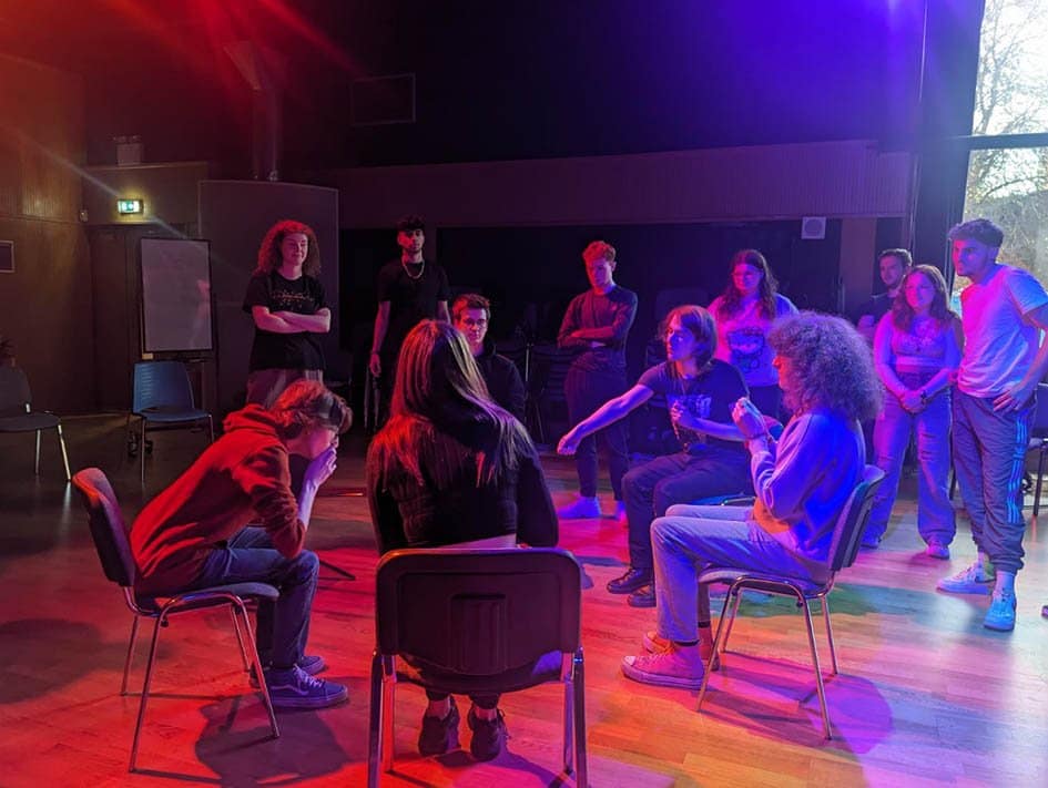 5 young people sit in a circle of chairs facing inwards towards one another on a wooden floor. Another group of 7 young people stand in the background outside the circle looking in on the people sitting down, some with crossed arms, some with arms by their side. The left side of the image is washed in a strong red light, while the right side of the image is washed in a strong blue/purple light.