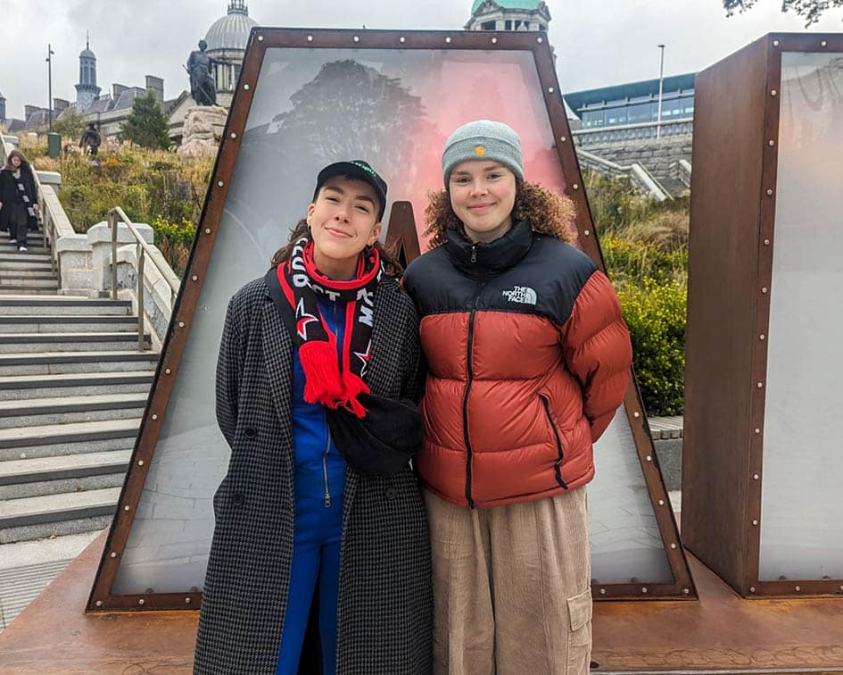 Two figures stand and smile at the camera, with a big letter 'A' in the background behind them. The figure on the left wears a long grey coat, a red and black scarf and a black cap. The figure on the right is wearing a brown and black puffer jacket and a grey beanie.